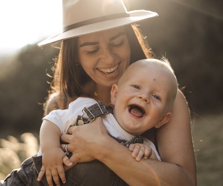 mother holding her baby on the arms and both laughing during sunset 
