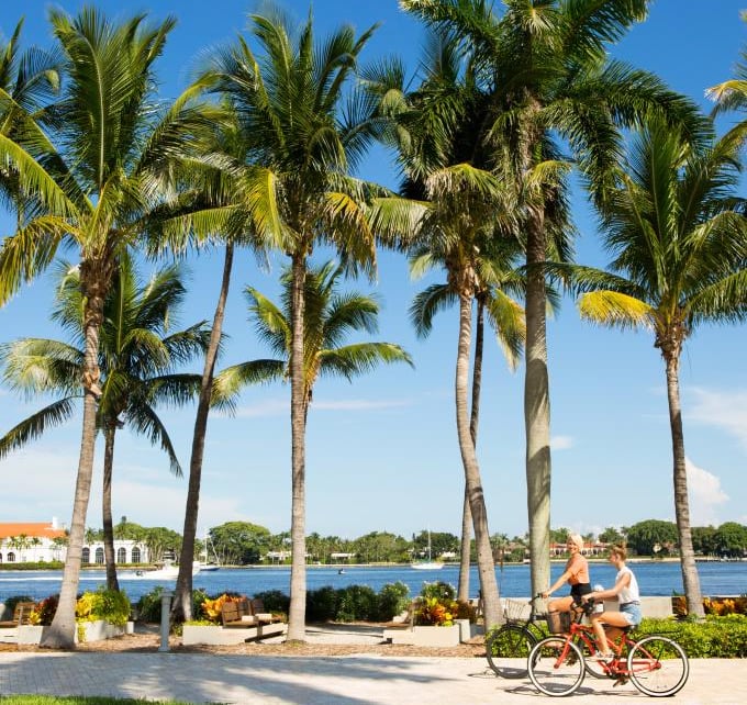 Two women cycling on a tropical waterfront path lined with palm trees in West Palm Beach, Florida.