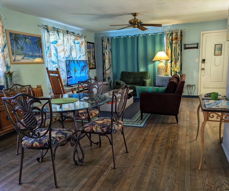 A tropical themed living and dining room with a glass table, dark hardwood floors, and teal curtains.