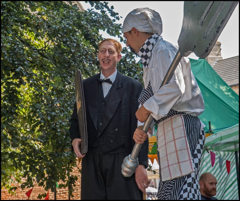 Street performers dressed as a comical chef and a formal butler act out a scene outdoors.