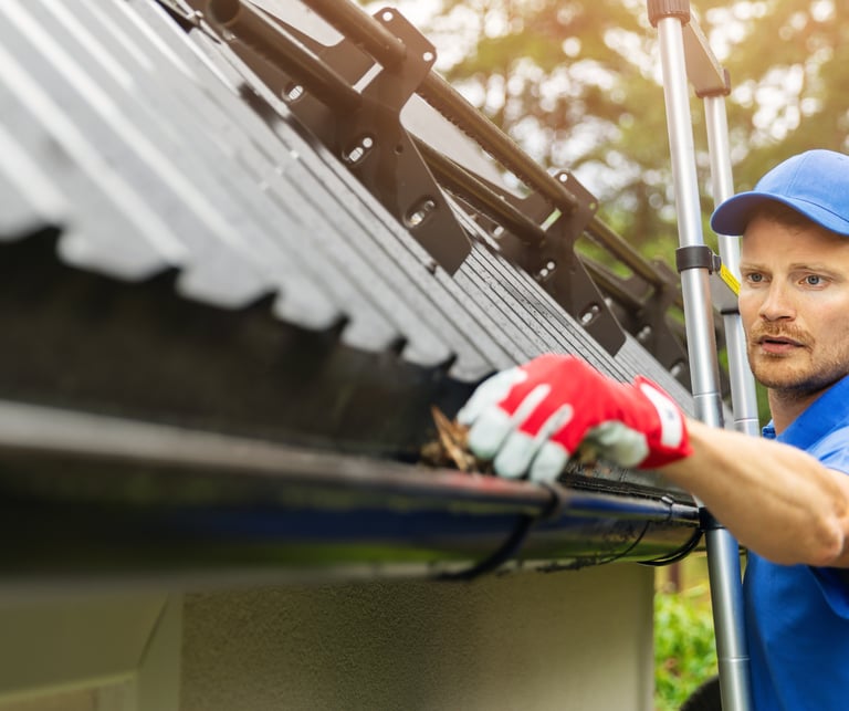 Professional contractor cleaning debris from a residential roof gutter system on a ladder.