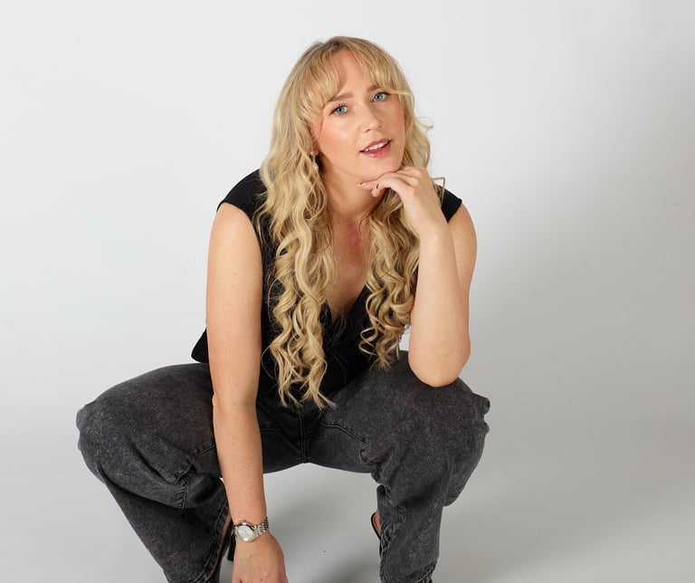 Blonde woman posing in a black sleeveless top and grey jeans with high heels on a white background.