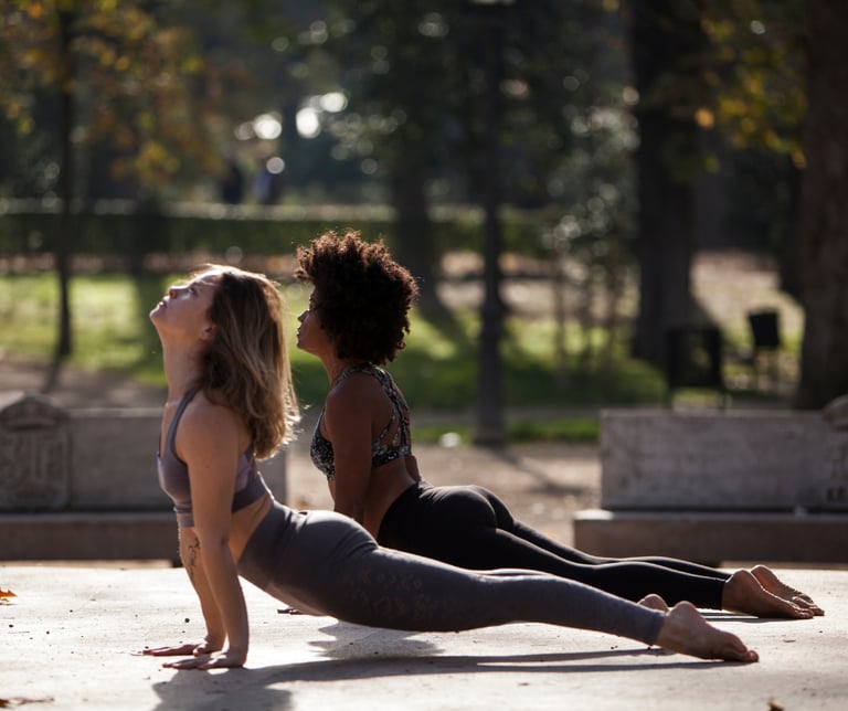 Two women practice upward facing dog yoga pose outdoors in a sunny park.