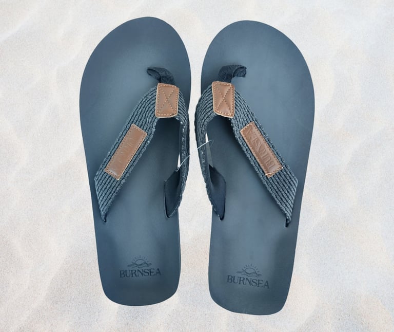 A pair of black Burnsea flip flops with grey fabric straps on a white sand background