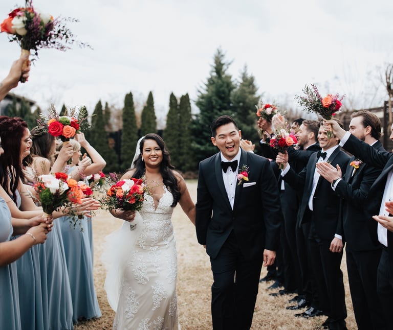 Smiling bride and groom walk through a cheering wedding party holding vibrant floral bouquets.