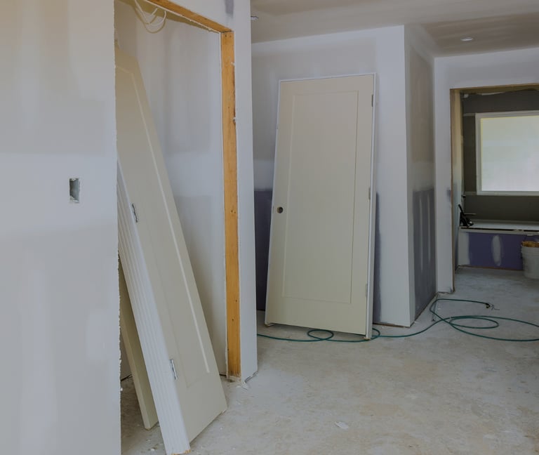 alt="Room in mid-renovation with drywall, exposed outlets, and interior door leaning against wall"