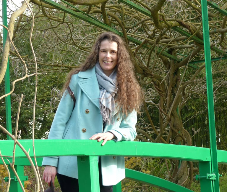 Elsa Errack, autrice - photo de l'auteure avec un manteau bleu sur un pont vert, entouré d'arbres.