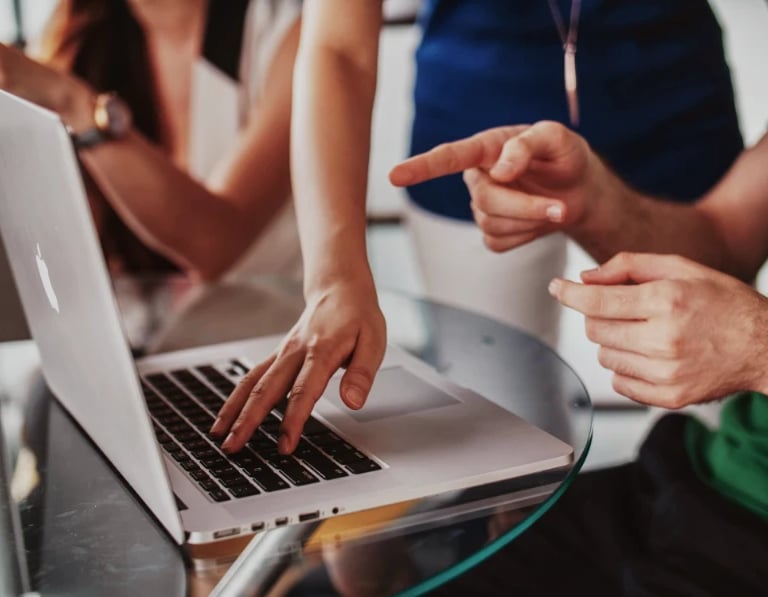 Personas colaborando en una mesa de cristal, señalando y escribiendo en un portátil.