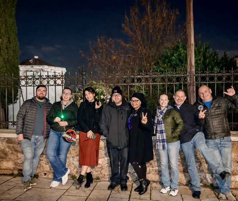 Group of eight tourists posing with peace signs and green flashlights in front of ornate iron fence during Athens Ghost Tour