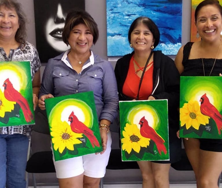 a group of smiling women holding up their paintings of a cardinal and a sunflower