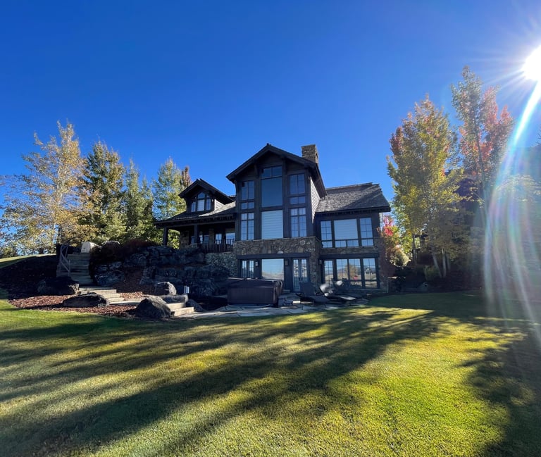 Luxury mountain estate with stone exterior, lush green lawn, and fall foliage under a bright sun.