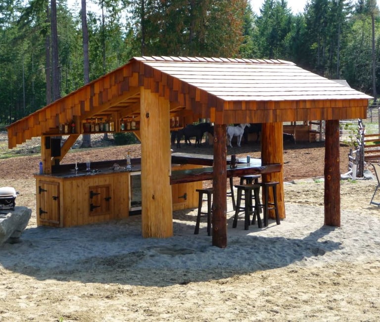 Rustic wooden outdoor tiki bar with stools, bottle shelves, and horses in the background.