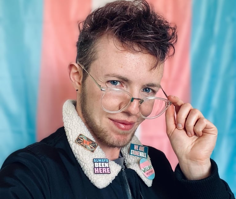 White trans masc in front of trans Pride flag wears a jacket with trans visibility pin badges