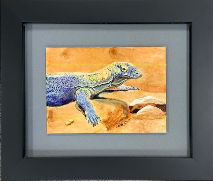 A framed ink painting of a blue and grey monitor lizard resting on orange sand.