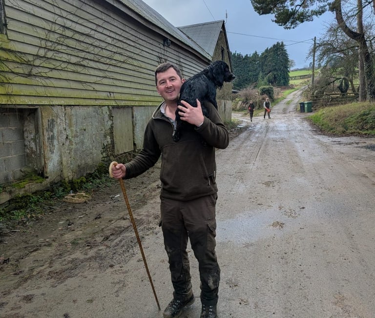 A smiling man in country attire carries a black spaniel dog on his shoulder along a rural farm track.
