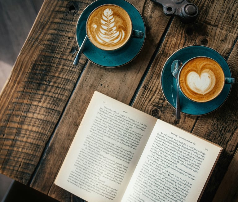 Two cups of latte art coffee sit on a rustic wooden table next to an open book in a cozy cafe.