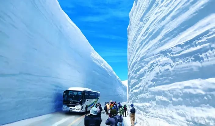 Tour bus driving through massive snow walls on a mountain road.