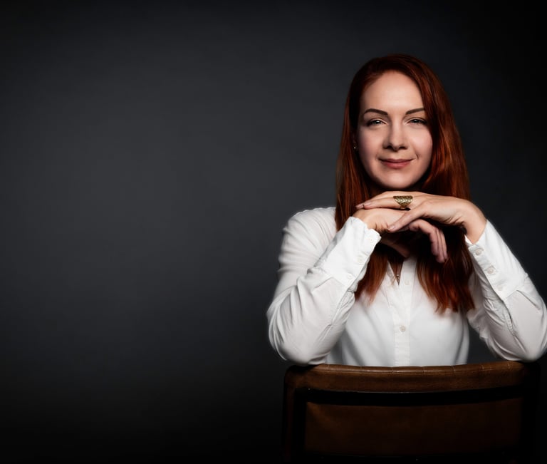 Professional corporate headshot of a smiling redhead woman in a white blouse on a dark background.