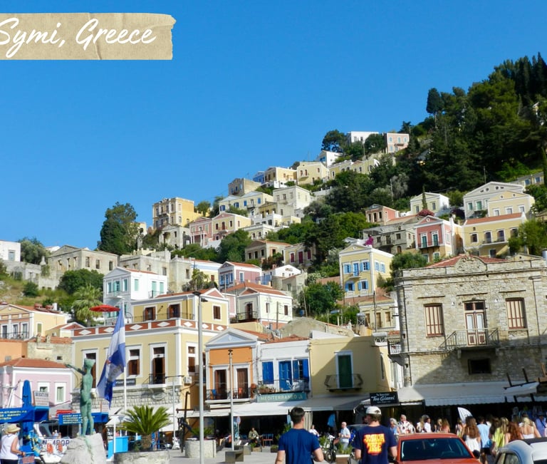 Colorful neoclassical houses on a hillside overlooking the harbor in Symi, Greece under a blue sky.