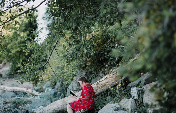 Person in red floral dress reading on a rock in a lush forest setting.