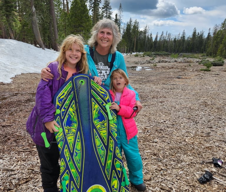 Teresa, Cecelia, and Mary Grace holding a sled by the snow and lake