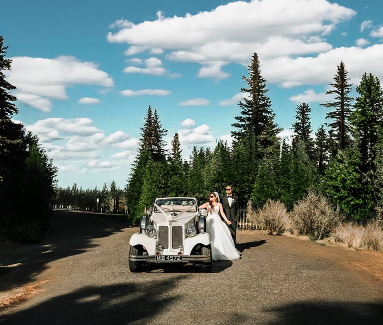 Bride and groom posing beside a vintage car on a scenic forest road, captured by Fred Art Studio.