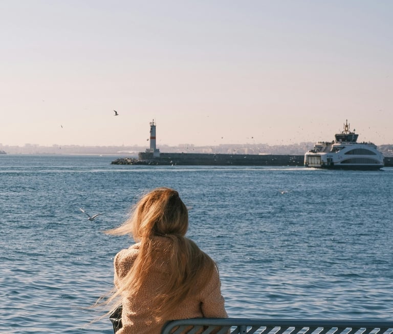 Donna seduta su una panchina al porto, di spalle, guarda il mare: vede traghetto e il faro