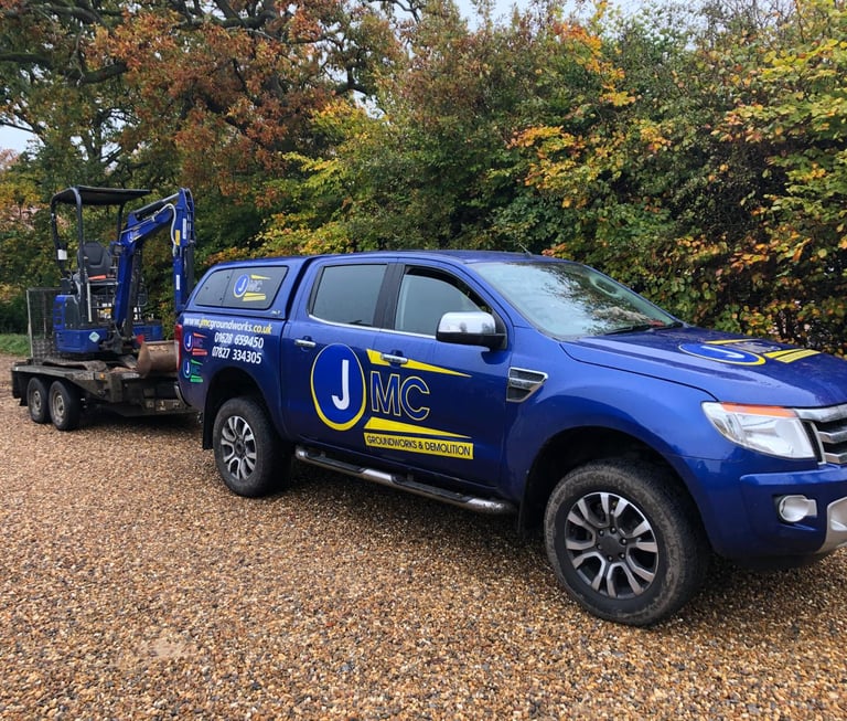 A blue JMC Groundworks Ford Ranger truck towing a mini excavator on a gravel driveway.