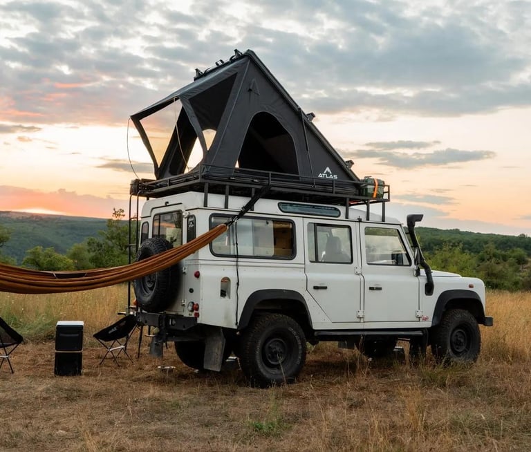 A white Land Rover Defender with a roof top tent and hammock parked in a field at sunset.
