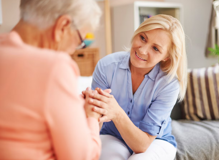 A blonde female caregiver provides compassionate home care while holding hands with an elderly woman.