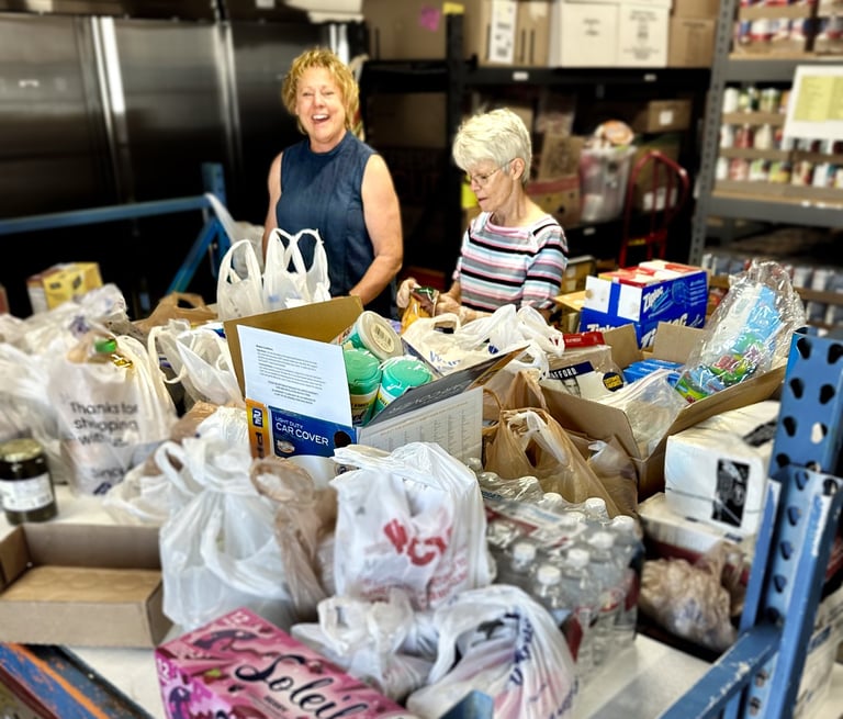 Two women standing behind table covered in bags of donated food in food pantry