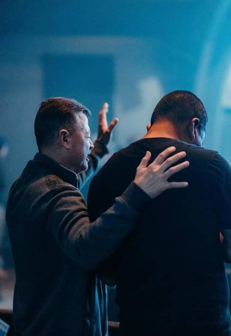 One person praying with a hand on another’s shoulder, symbolizing support, prayer, and community.