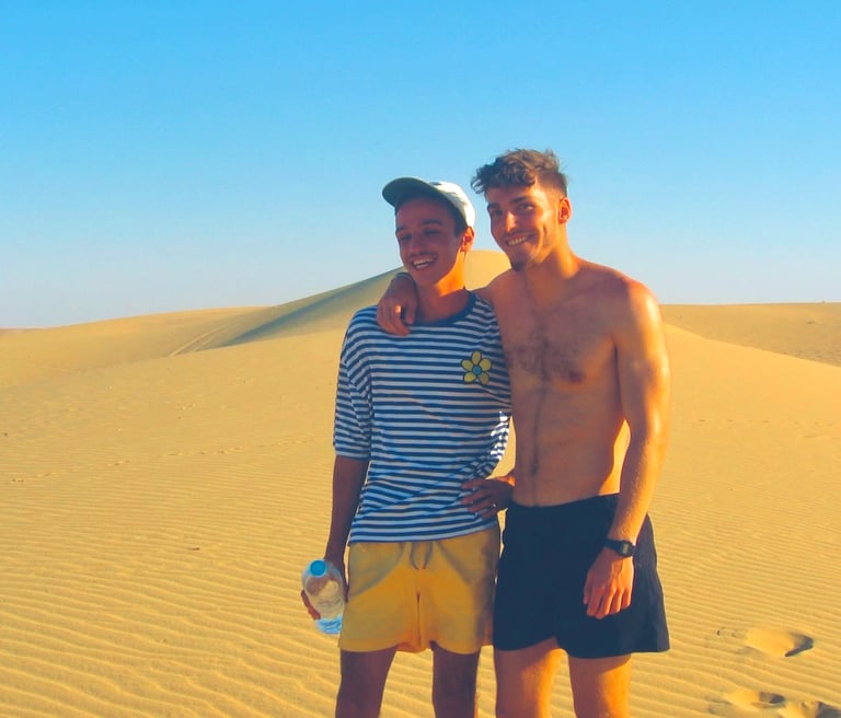  Two young men, smiling and embracing, stand in a desert landscape. The man on the left wears a stri
