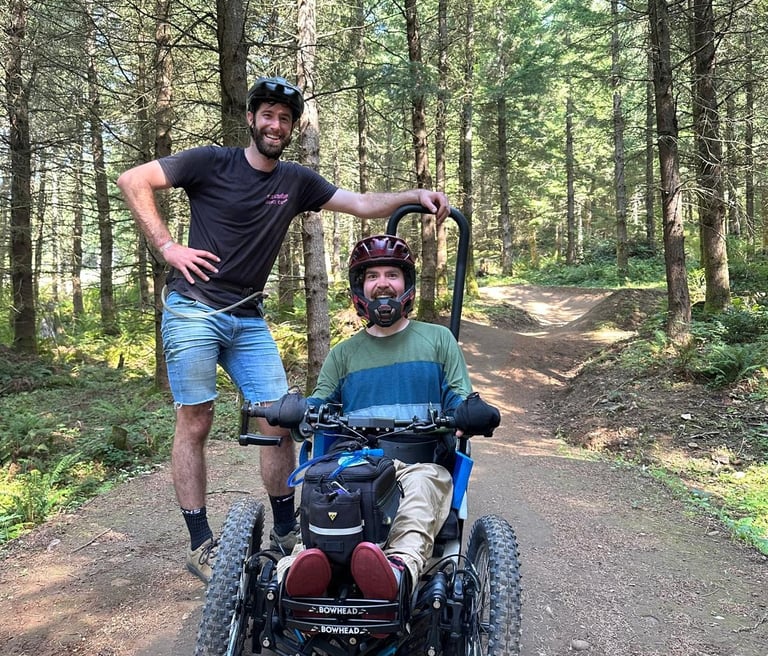 Sean seated in his 3-wheeled adaptive mountain bike on a forest trail with his friend Alex standing