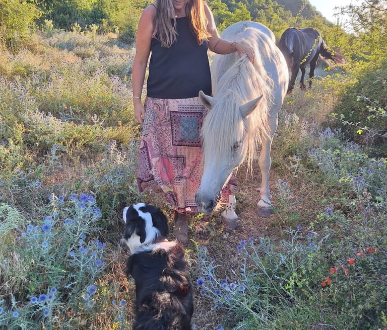 Vrouw staat naast een wit paard in een weiland met bloemen, ze legt haar hand op het paard
