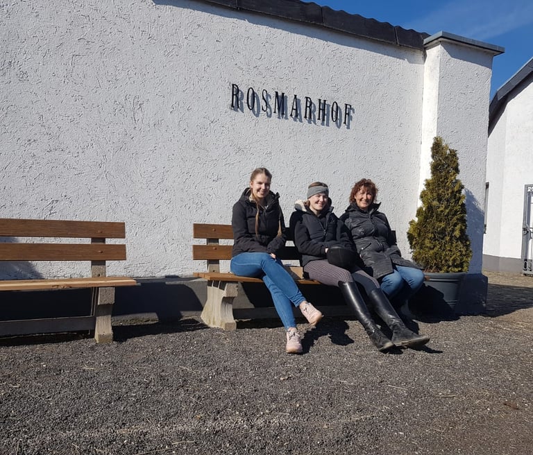 Three women sitting on a bench outside the Rosmarhof equestrian facility on a sunny day.