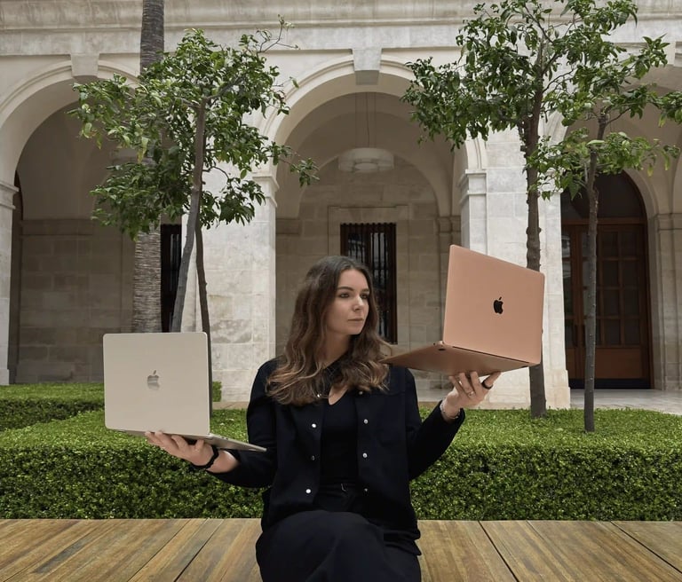 A woman balances two Apple MacBooks while sitting in a stone courtyard, illustrating remote work technology.