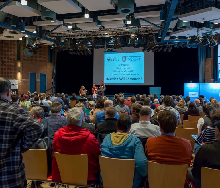 Viele Menschen sitzen im Publikum und hören den Redner*innen auf der Bühne zu.