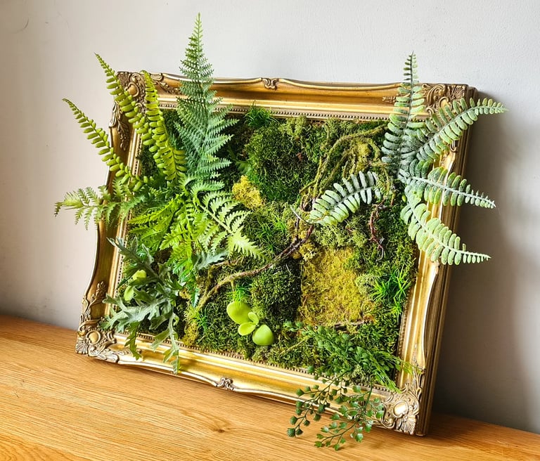 Preserved moss wall art with green ferns in an ornate gold vintage picture frame on a wooden table.