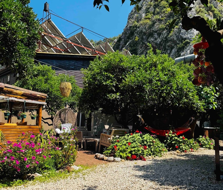 Olympos Kakule Evleri’nin bahçesi; çiçeklerle süslenmiş oturma alanı, hamak ve yemyeşil ağaçlar.