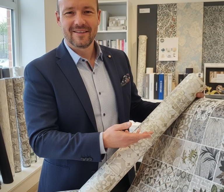 Professional interior designer presenting a roll of floral and geometric patterned wallpaper in a showroom.