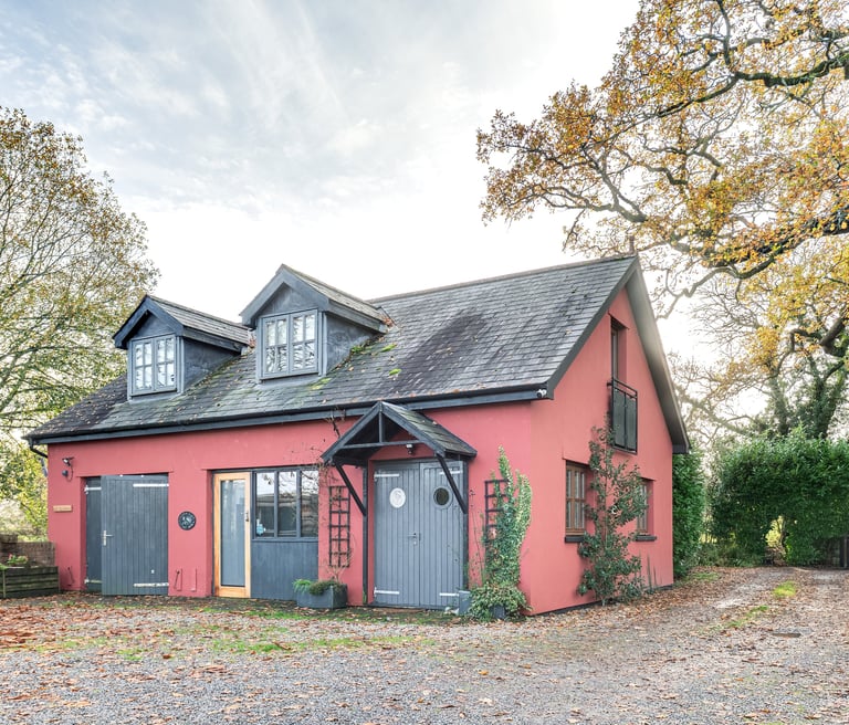Red country cottage with grey barn doors and dormer windows surrounded by autumn trees.