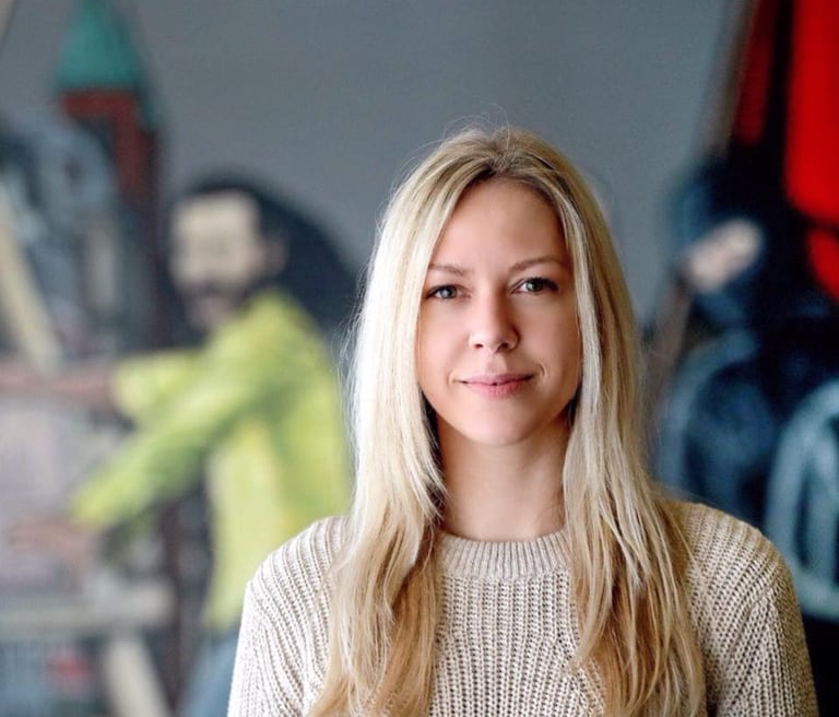 Rikke Lydeking in a beige knit sweater smiling in an art studio with paintings in the background.