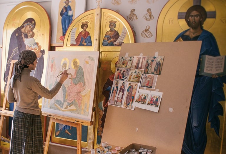 An artist paints a religious icon on an easel inside a studio filled with Byzantine art.