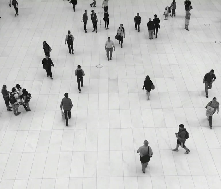 A high-angle black and white photo of people walking across a large, white tiled floor in a public space.
