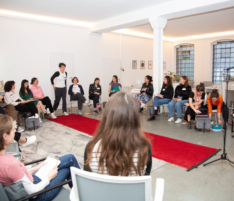Workshop room, with red carpet in middle, women sitting around it, 1 woman walking down, towards mic