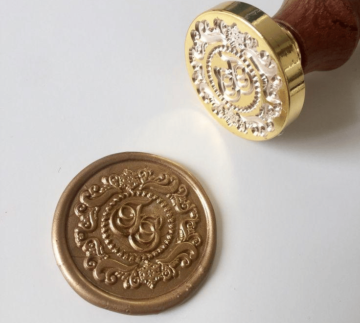 Custom brass wax seal stamp and a finished gold wax seal with an ornate monogram design on white paper.