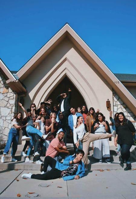 Energetic group striking playful poses in front of a church-like building under a bright blue sky.