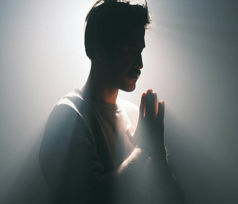 Silhouetted figure with hands pressed in prayer, surrounded by radiant light against a dark backgrou