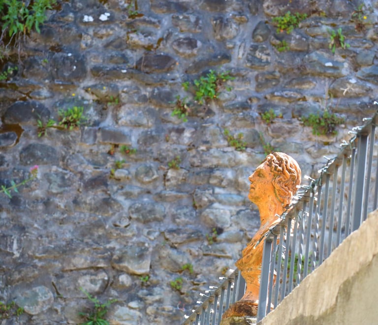 A classical terracotta bust sculpture on a wrought iron railing against an ancient stone wall background.